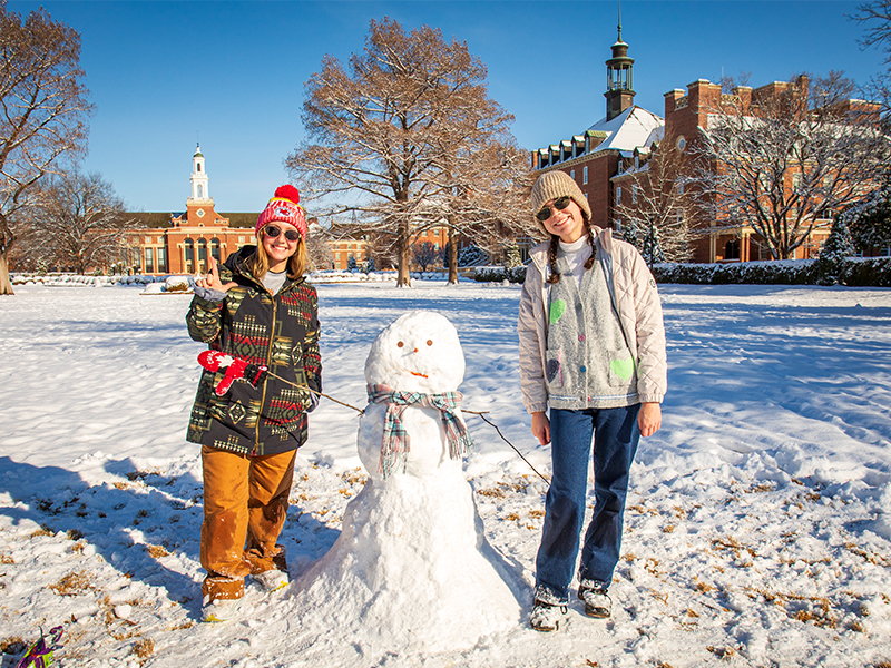 Two students standing next to a snowman they made with Edmon Low Library in the background.